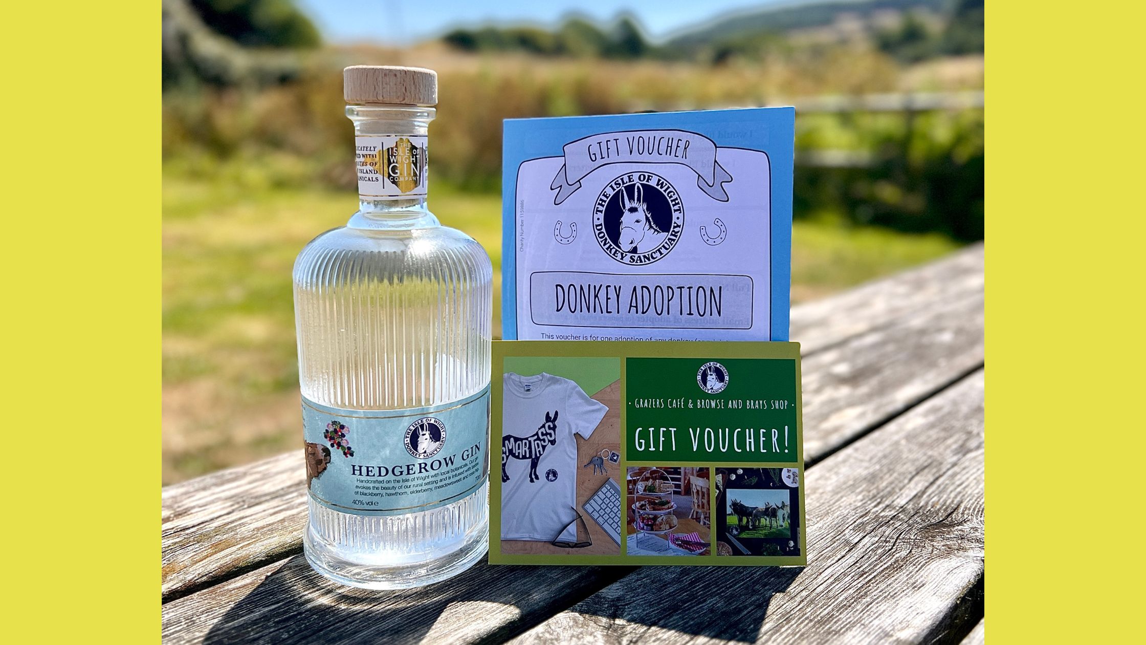 a bottle of gin and two gift vouchers on a wooden table with grass and blue skies in the background